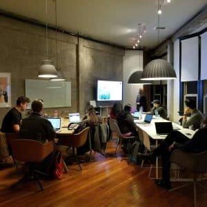 A group of people is sitting around tables in a modern office setting, working on laptops and other devices. A person stands at the front near a large screen displaying information about ETL in Marketing. The room has a polished wood floor, pendant lights, and large windows with shades.