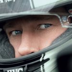 Close-up of a person wearing a racing helmet with the brand "Bell" visible. The person's intense eyes are looking out from the helmet's visor, and they are dressed in racing gear with a partially visible "Shark" logo. The image feels like an epic trailer for an F1 movie, conveying focus and determination.