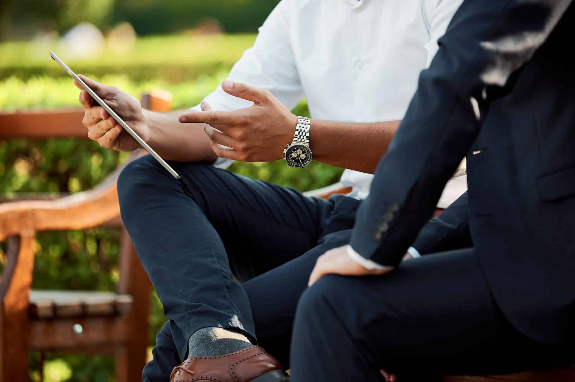 Two people are sitting on a bench outdoors, one wearing a white shirt and showing something on a tablet to the other, who is dressed in a dark suit. Their faces are not visible, but they appear to be engaged in a discussion about Google Ads strategy.