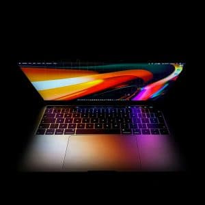 A laptop with a colorful and bright abstract wallpaper on its screen. The laptop is surrounded by darkness, highlighting the keyboard and the screen's vivid colors and patterns. The image showcases the device in a dimly lit environment, possibly featuring the latest mac Sonoma 14.5 update.