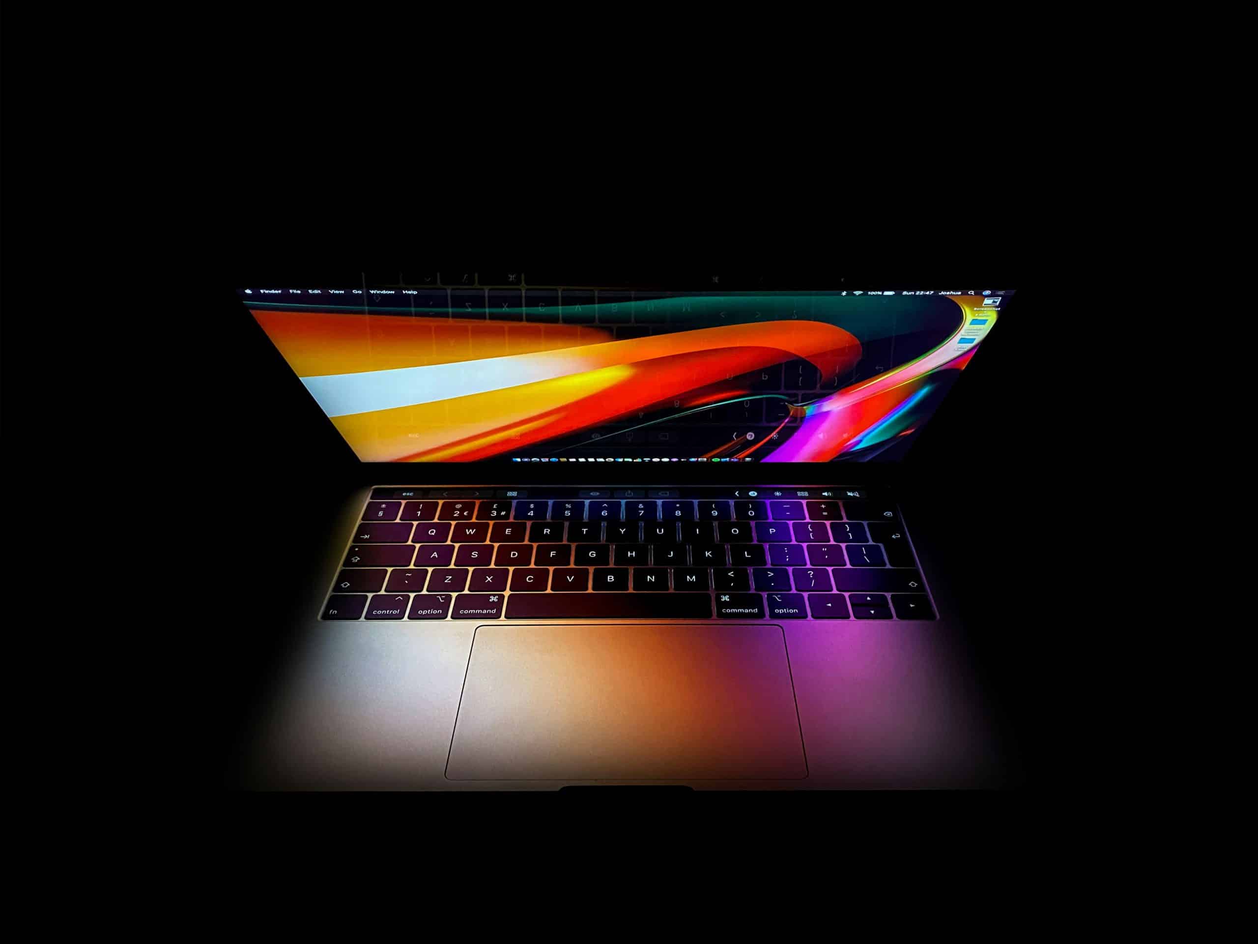 A laptop with a colorful and bright abstract wallpaper on its screen. The laptop is surrounded by darkness, highlighting the keyboard and the screen's vivid colors and patterns. The image showcases the device in a dimly lit environment, possibly featuring the latest mac Sonoma 14.5 update.