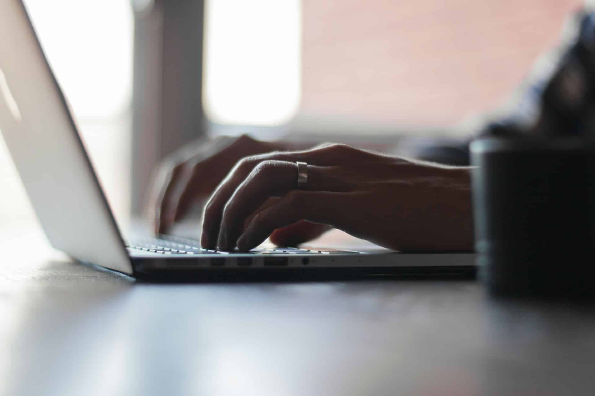 Close-up of hands typing on a laptop keyboard with natural light streaming through a window in the background. The person, who appears to be working on Google Ads, is wearing a ring on their ring finger while using the laptop. A blurred cup is visible in the foreground on the right side.