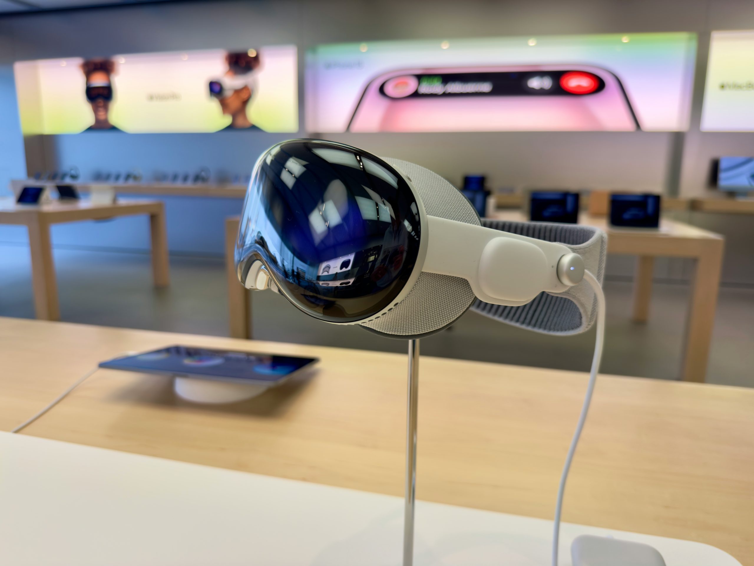 A sleek VR headset, reminiscent of the Apple Vision Pro, is displayed on a stand in a modern electronics store. The headset has a reflective visor and an adjustable strap. In the background, screens and tablets are set up on wooden tables, with illuminated digital displays and advertisements on the walls.