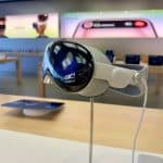 Close-up of a virtual reality headset on display in a tech store. The reflective visor shows the store's interior. In the background, digital screens and tables with various tech devices are visible, showcasing US Vision Pro sales amid IDC reports of a recent decline in the market.