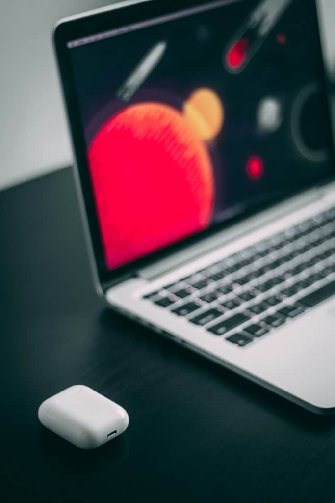 A laptop on a dark table, its screen displaying abstract red and yellow graphics reminiscent of Playlist Artwork, is partially visible. In front of the laptop, there is a white wireless earbud case | M4 Mac Models.