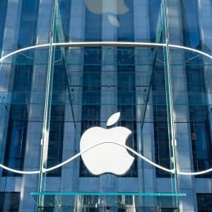 A glass building entrance with a large white Apple logo in the center. Surrounding the logo is a thin, white neon light outline resembling the shape of a Vision Pro headset. The reflections on the glass show surrounding architecture, creating a sleek, cutting-edge visual that blends Apple's signature style with advanced tech aesthetics.