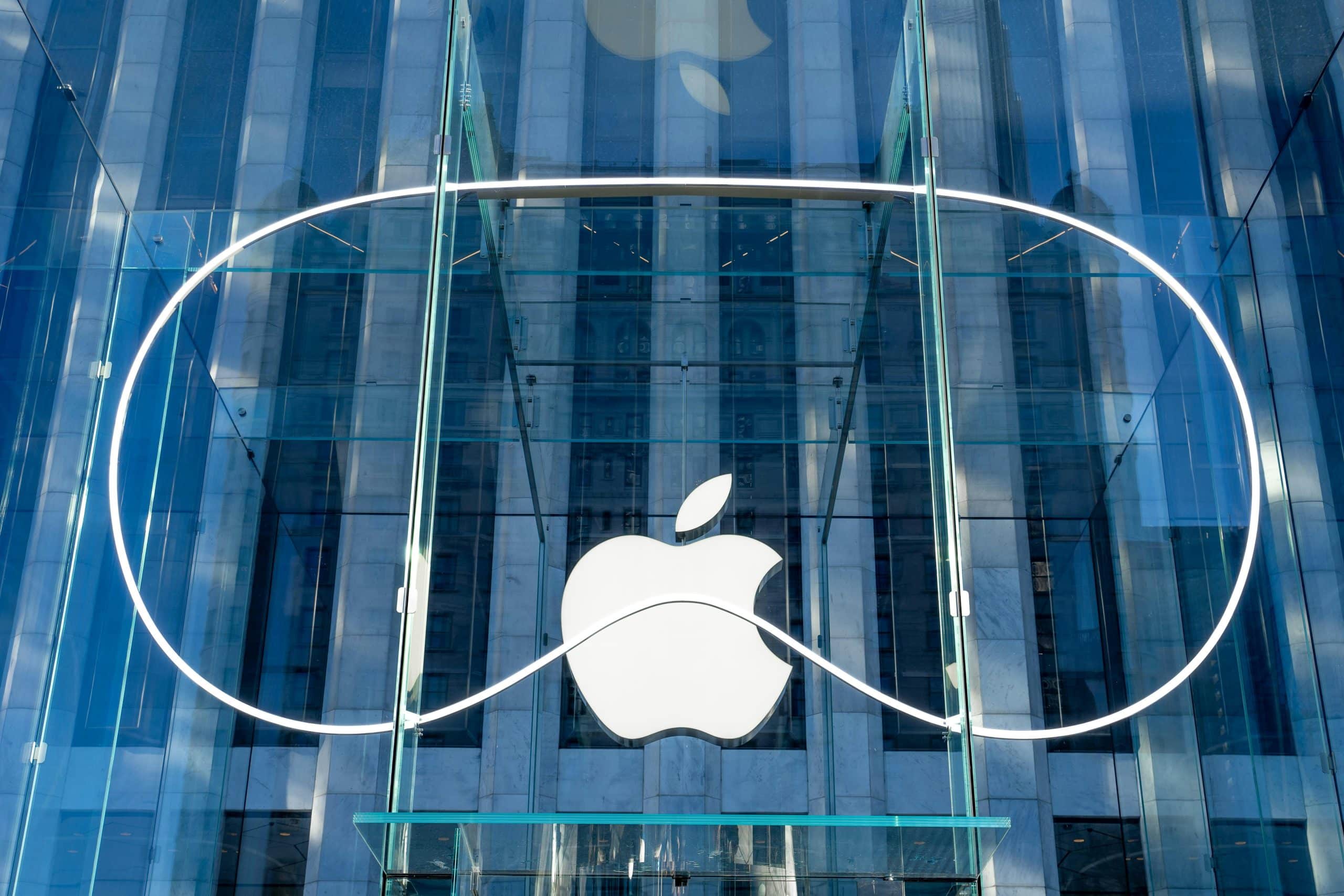 A glass building entrance with a large white Apple logo in the center. Surrounding the logo is a thin, white neon light outline resembling the shape of a Vision Pro headset. The reflections on the glass show surrounding architecture, creating a sleek, cutting-edge visual that blends Apple's signature style with advanced tech aesthetics.