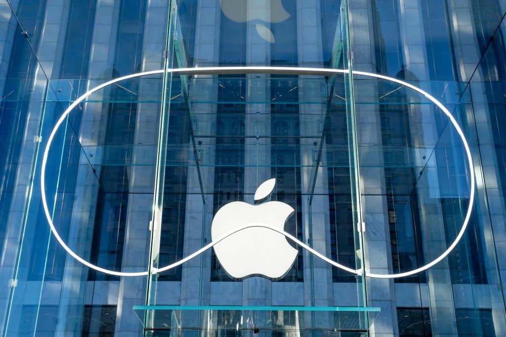 The image shows a large Apple logo displayed at the entrance of an Apple Store. The logo is mounted on a glass facade with a sleek, modern design. Amid reports of declining 75% US Vision Pro sales, the store's glass exterior reflects nearby buildings, enhancing its urban setting.