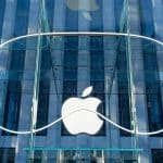 The image shows a large Apple logo displayed at the entrance of an Apple Store. The logo is mounted on a glass facade with a sleek, modern design. Amid reports of declining 75% US Vision Pro sales, the store's glass exterior reflects nearby buildings, enhancing its urban setting.