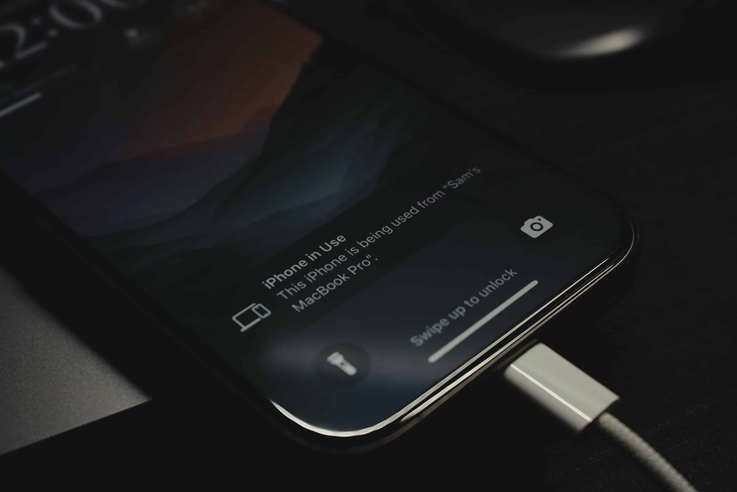 Close-up of a smartphone screen displaying a dark background with a notification. The lower part of the screen shows "iPhone in Use" and a message about sharing its usage from a MacBook Pro. The phone, rumored to be the iPhone 16 Pro with faster charging speeds, is connected to a charger.