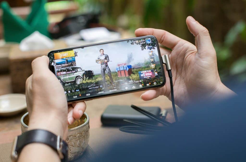 A person is holding an Apple smartphone horizontally with both hands, playing a video game on iOS. The game on the screen features a character selection interface with the "START" button visible. Various gaming equipment and blurred outdoor elements are in the background.