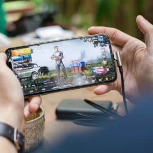 A person is holding an Apple smartphone horizontally with both hands, playing a video game on iOS. The game on the screen features a character selection interface with the "START" button visible. Various gaming equipment and blurred outdoor elements are in the background.