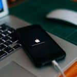 A smartphone displaying the Apple logo with a loading bar, potentially updating to iOS 18, is connected to a laptop with a grey keyboard. The laptop rests on a green cutting mat, and a white computer mouse is placed nearby on the wooden surface.