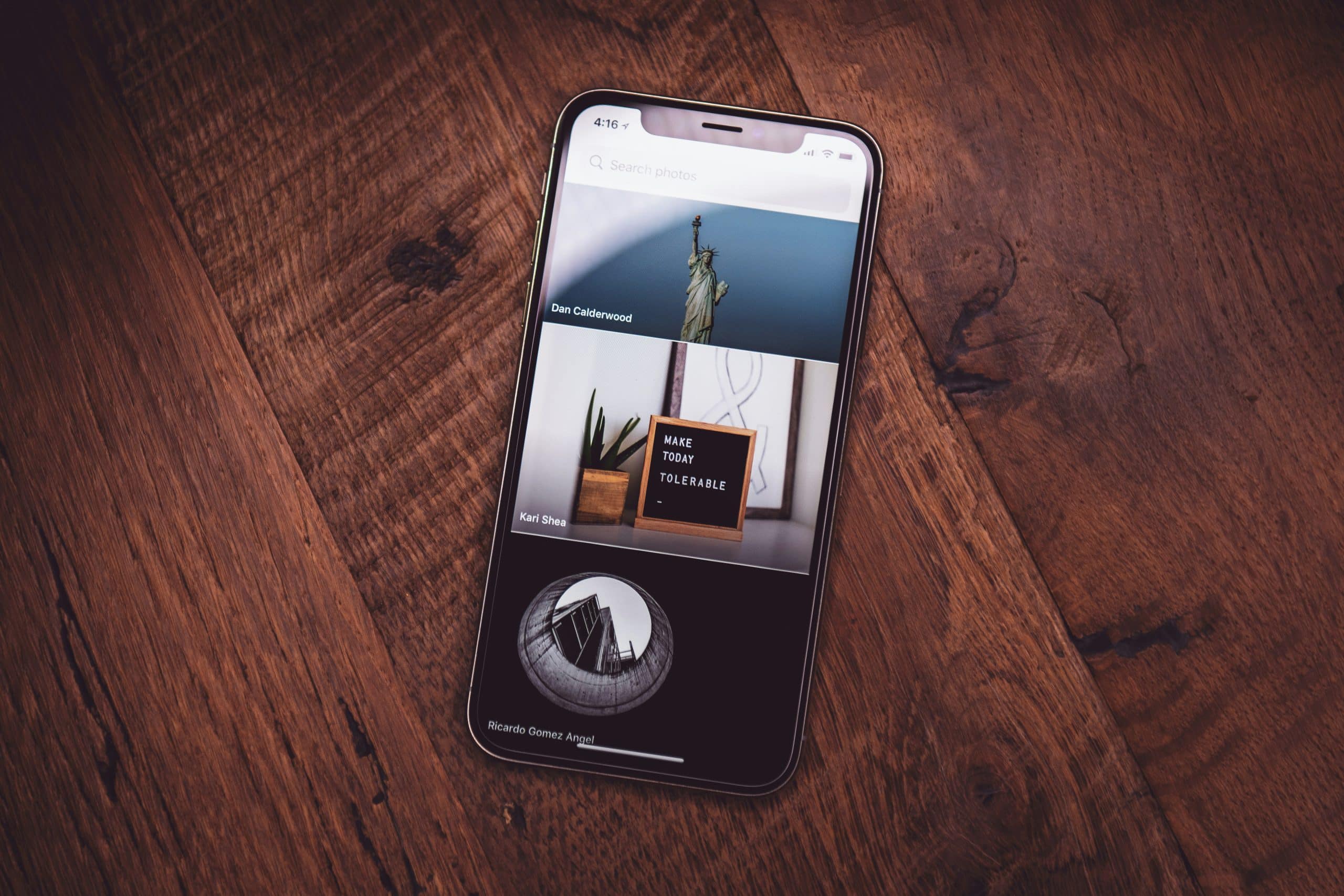 A sleek Apple smartphone displaying a gallery app lies on a wooden surface. The screen shows three photos: the Statue of Liberty, a framed quote saying "MAKE TODAY TOLERABLE" near some plants, and a black and white image of modern architecture, as if from a carefully recovered album.