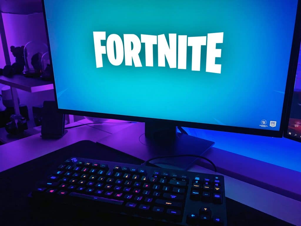 A computer screen displays the "Fortnite" game logo against a blue background. In front of the screen, an RGB-lit keyboard is visible on a desk, next to which lie two sleek iPhones. The setup is illuminated with purple and blue lighting.