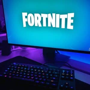 A computer screen displays the "Fortnite" game logo against a blue background. In front of the screen, an RGB-lit keyboard is visible on a desk, next to which lie two sleek iPhones. The setup is illuminated with purple and blue lighting.