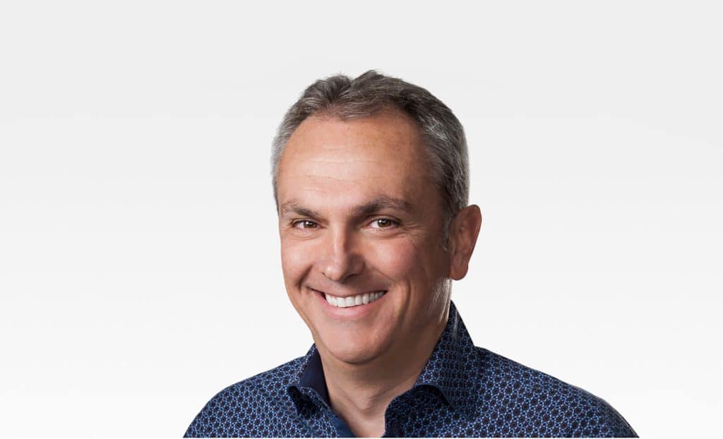 A middle-aged man with short, graying hair smiles at the camera. He is wearing a dark blue patterned shirt. The background is plain and white, giving the image a professional, corporate feel akin to an Apple CFO portrait.