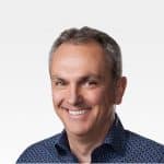 A middle-aged man with short, graying hair smiles at the camera. He is wearing a dark blue patterned shirt. The background is plain and white, giving the image a professional, corporate feel akin to an Apple CFO portrait.