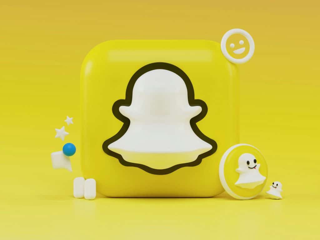 A 3D rendering of the Snapchat logo. The image features a white ghost outline on a yellow square background with various related 3D elements, including stars, emojis, and speech bubbles, surrounding the logo. The entire scene captures the playful and engaging spirit of Snapchat.