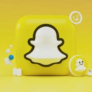 A 3D rendering of the Snapchat logo. The image features a white ghost outline on a yellow square background with various related 3D elements, including stars, emojis, and speech bubbles, surrounding the logo. The entire scene captures the playful and engaging spirit of Snapchat.