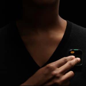 A person against a dark background is pinching a small, square black device with a corner display and an orange button. The Humane AI Pin, held near the chest area of their black sweater, represents the cutting-edge technology highlighted in the new report.