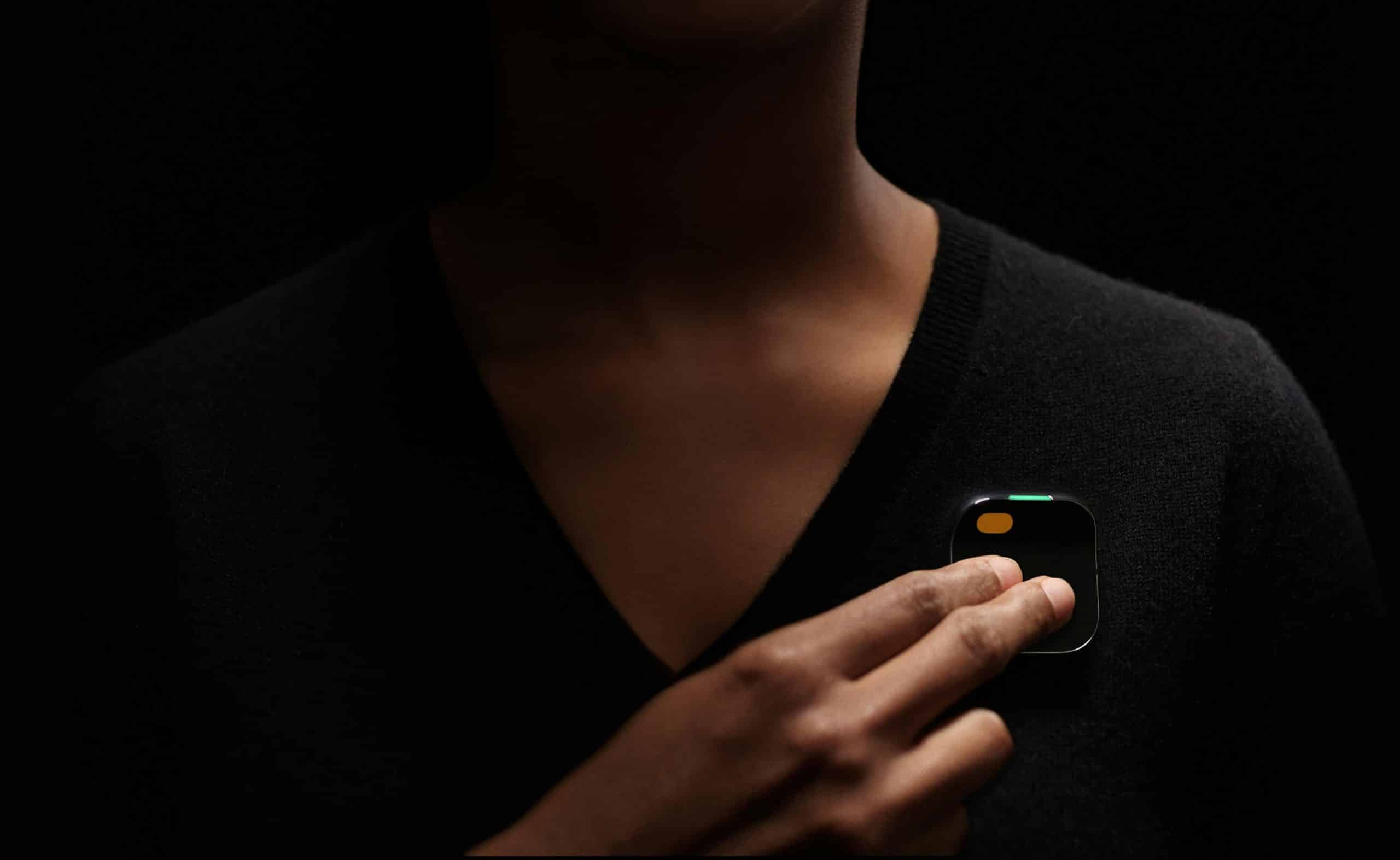 A person against a dark background is pinching a small, square black device with a corner display and an orange button. The Humane AI Pin, held near the chest area of their black sweater, represents the cutting-edge technology highlighted in the new report.