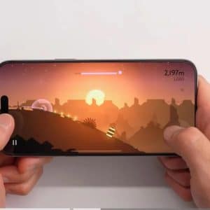 Hands hold a smartphone horizontally with a game displayed on the screen. The game features a silhouette landscape with a sunset, with the player character moving through the terrain. Various game elements like score and progress bar are visible.