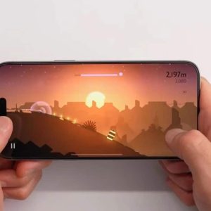 Hands hold a smartphone horizontally with a game displayed on the screen. The game features a silhouette landscape with a sunset, with the player character moving through the terrain. Various game elements like score and progress bar are visible.