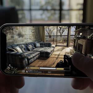 A person holds a smartphone horizontally, displaying a video game that features an in-game view of a living room with a plaid sofa, wooden coffee table, fireplace, and large window. An Apple logo is visible in the bottom right corner of the image, highlighting one of Germany's popular iPhone apps.