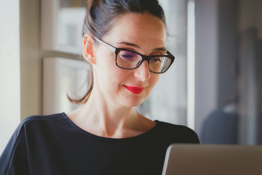 A person with dark hair tied back, wearing black-framed glasses and a black top, smiles while looking at a laptop in front of them. They are inside a well-lit room with a large window in the background, possibly researching responsible procurement practices.