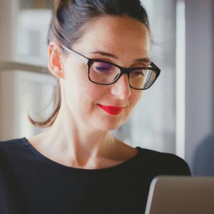 A person with dark hair tied back, wearing black-framed glasses and a black top, smiles while looking at a laptop in front of them. They are inside a well-lit room with a large window in the background, possibly researching responsible procurement practices.