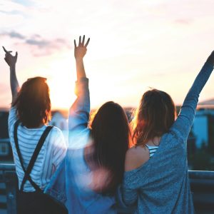 Three people stand on a bridge at sunset, facing away from the camera. They have their hands raised, with two making peace signs. Dressed casually in shirts and jackets, they enjoy a meaningful conversation while taking in the scenic, sunlit view.