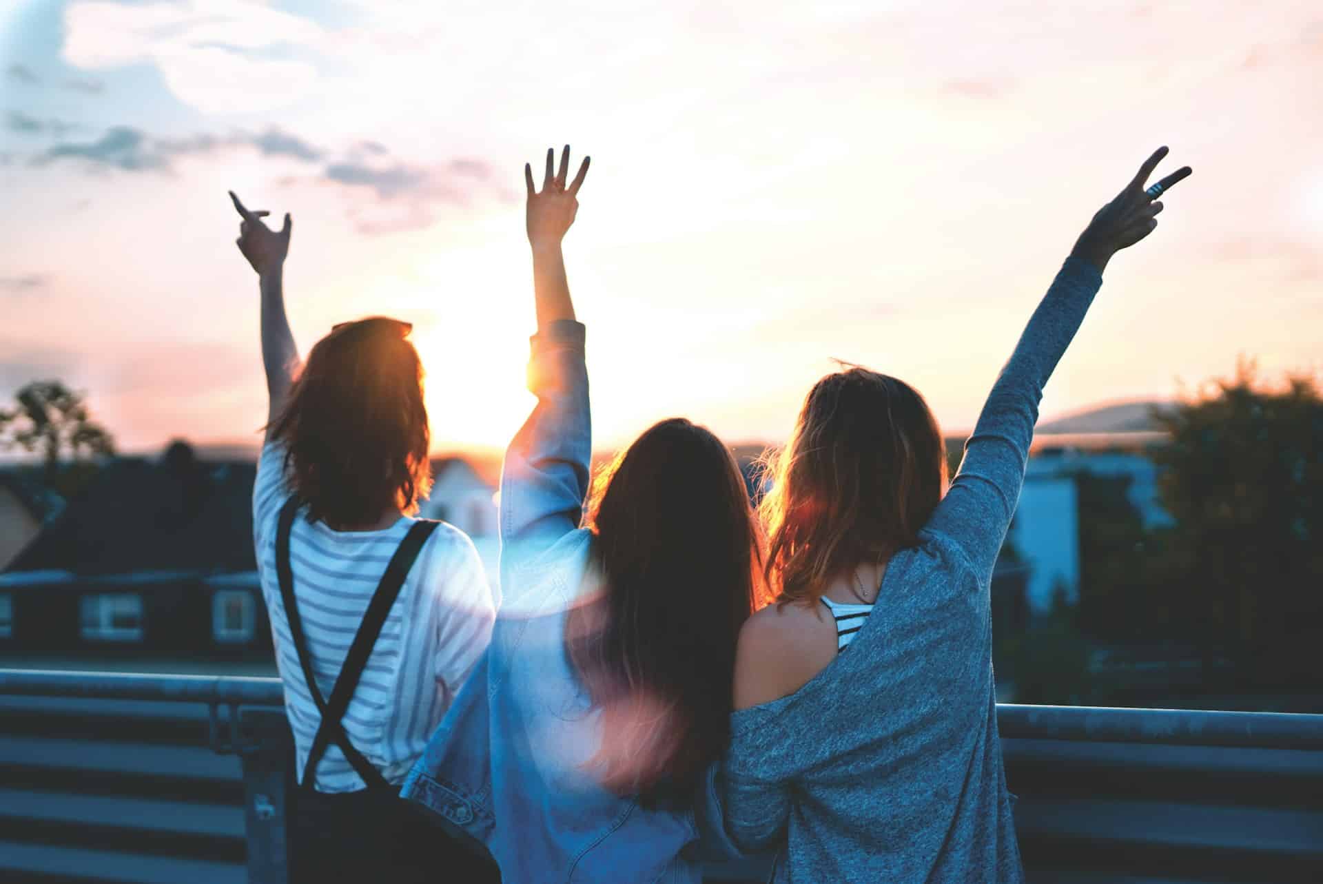 Three people stand on a bridge at sunset, facing away from the camera. They have their hands raised, with two making peace signs. Dressed casually in shirts and jackets, they enjoy a meaningful conversation while taking in the scenic, sunlit view.