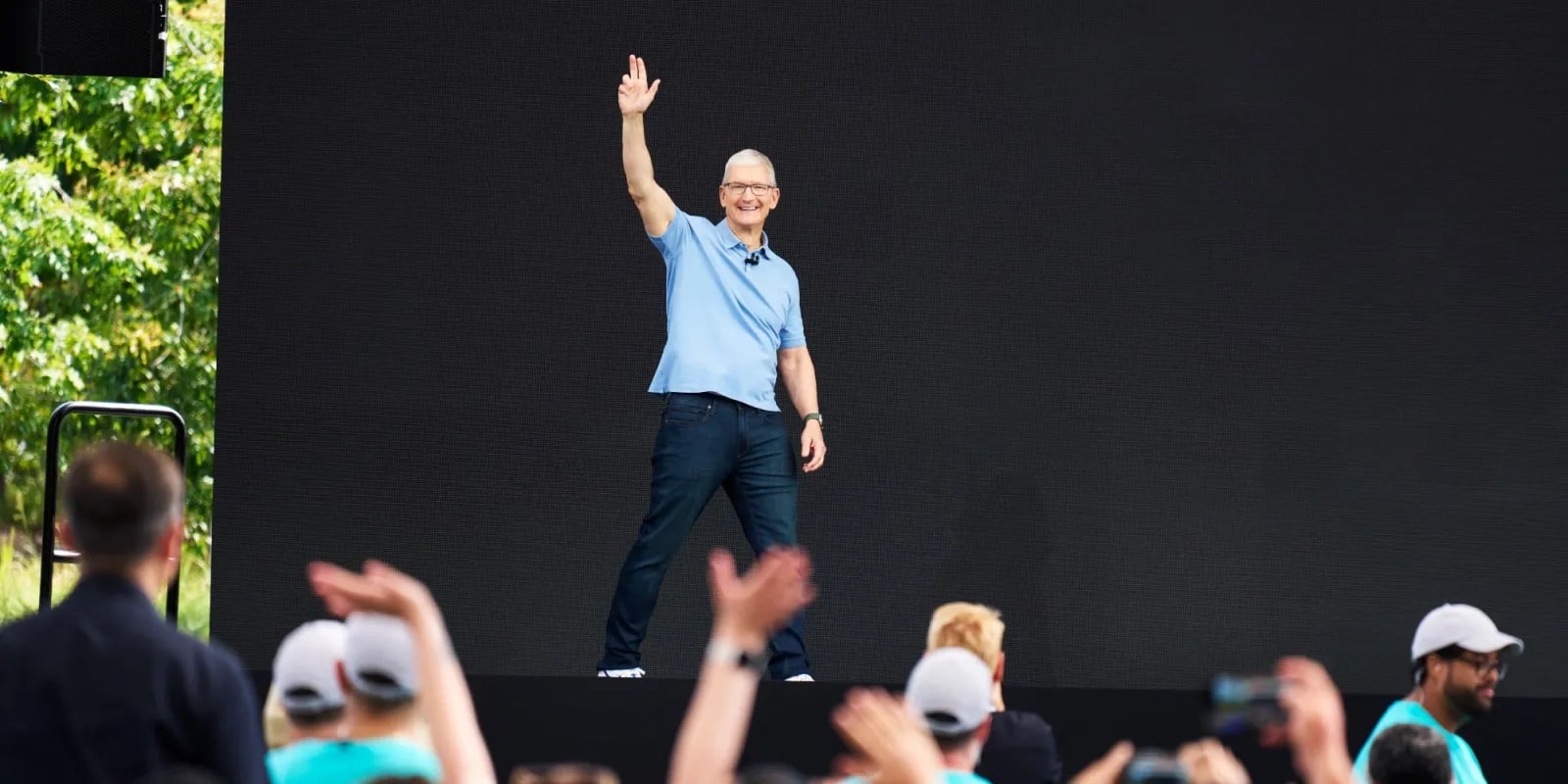 A person wearing glasses, a light blue polo shirt, and jeans is standing on a stage, waving with one hand raised. They are smiling, as people in the foreground clap and raise their hands in celebration of Apple's impressive 3Q earnings report. The background is black.