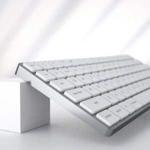 A sleek, white computer keyboard is angled upwards, with the front end elevated by a small white cube on a clean, white surface. Sunlight casts soft shadows across the scene, creating a minimalist and modern aesthetic that perfectly complements Mac users' preference for seamless keyboard integration.