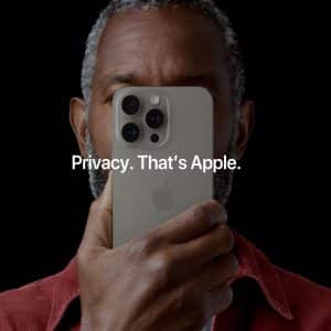 A person holds an iPhone in front of their face, almost covering it completely. The text overlay on the image reads, "Privacy. That's Apple." The background is black, and the person wears a red shirt. The iPhone has a light-colored case and three camera lenses.