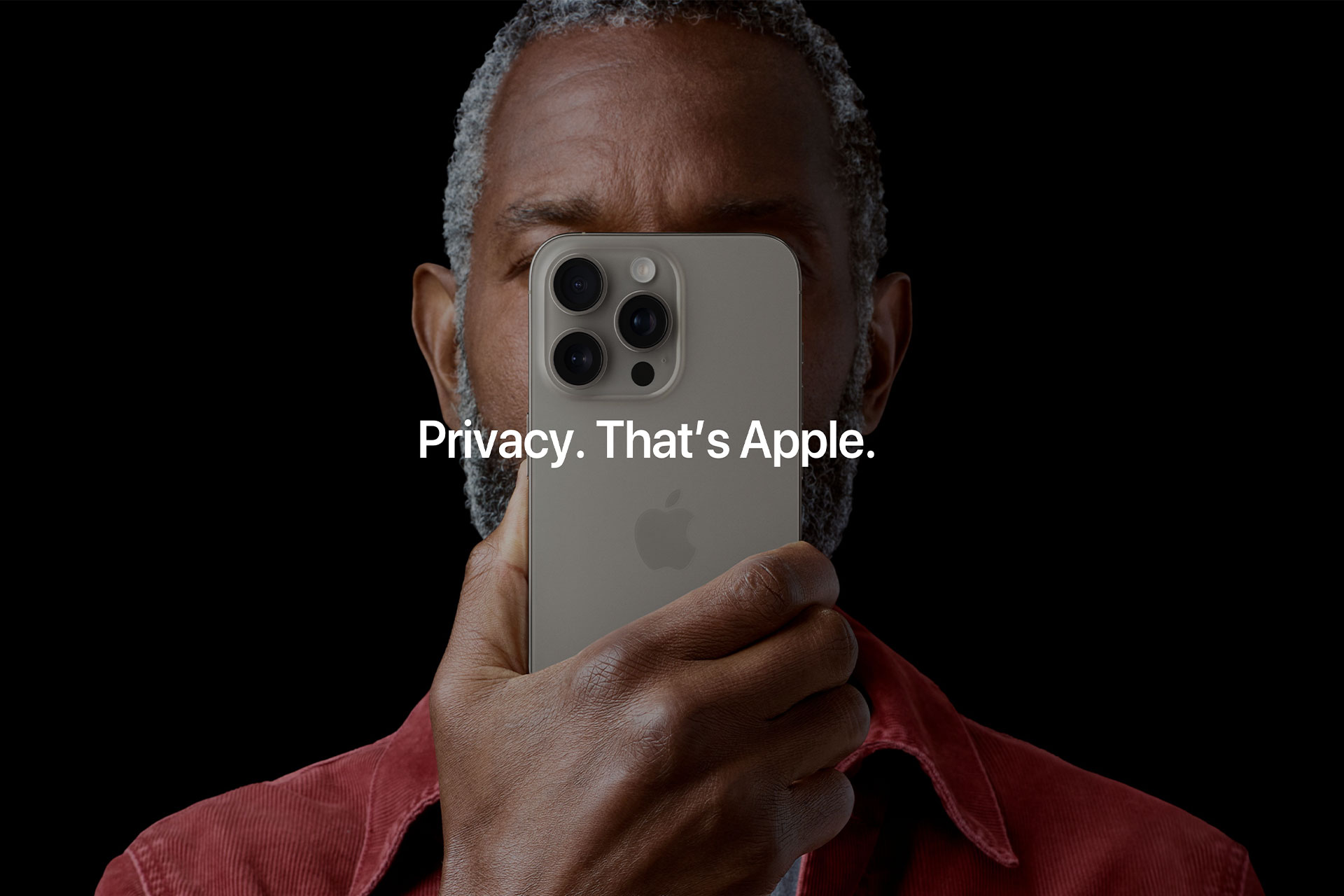 A person holds an iPhone in front of their face, almost covering it completely. The text overlay on the image reads, "Privacy. That's Apple." The background is black, and the person wears a red shirt. The iPhone has a light-colored case and three camera lenses.