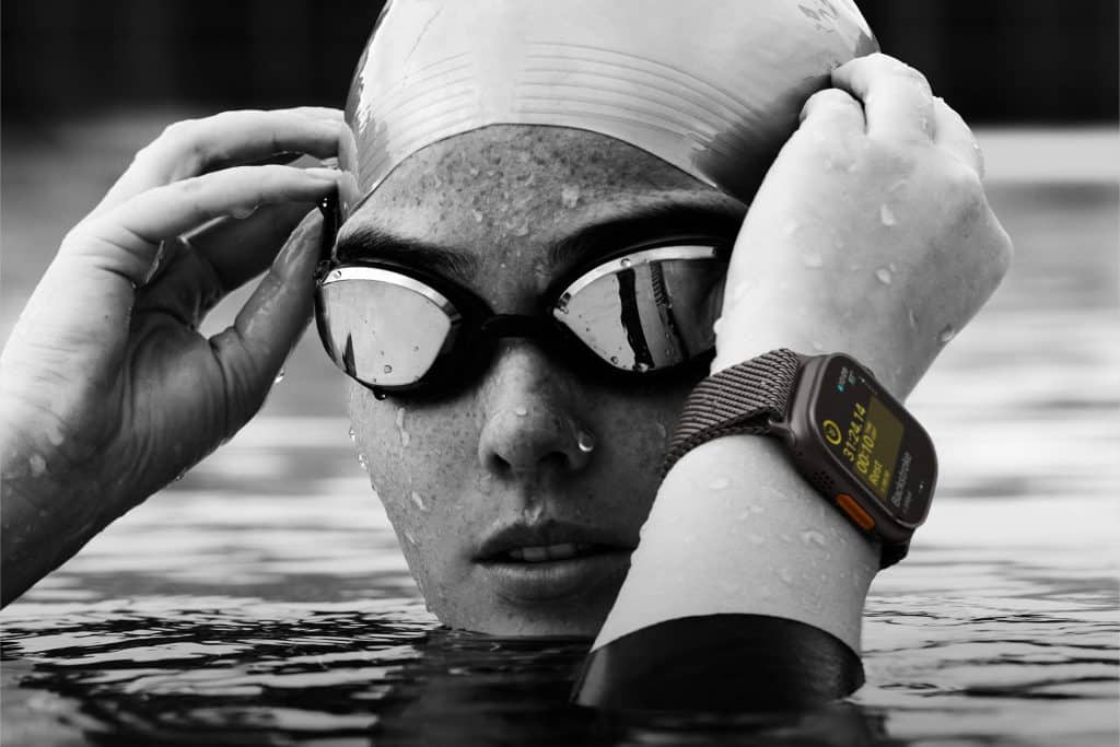 Live Activities 11: A swimmer wearing a swim cap and goggles adjusts their goggles while partially submerged in water. An advanced smartwatch with a detailed, visible display is on their wrist, indicating they are likely tracking their swimming performance or other metrics.