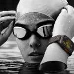 Live Activities 11: A swimmer wearing a swim cap and goggles adjusts their goggles while partially submerged in water. An advanced smartwatch with a detailed, visible display is on their wrist, indicating they are likely tracking their swimming performance or other metrics.