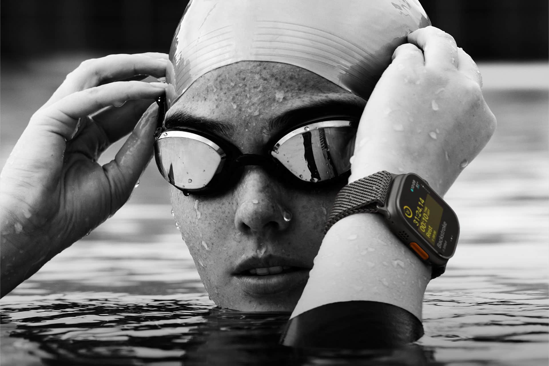 A swimmer wearing a swim cap and goggles adjusts their goggles while partially submerged in water. An advanced smartwatch with a detailed, visible display is on their wrist, indicating they are likely tracking their swimming performance or other metrics.