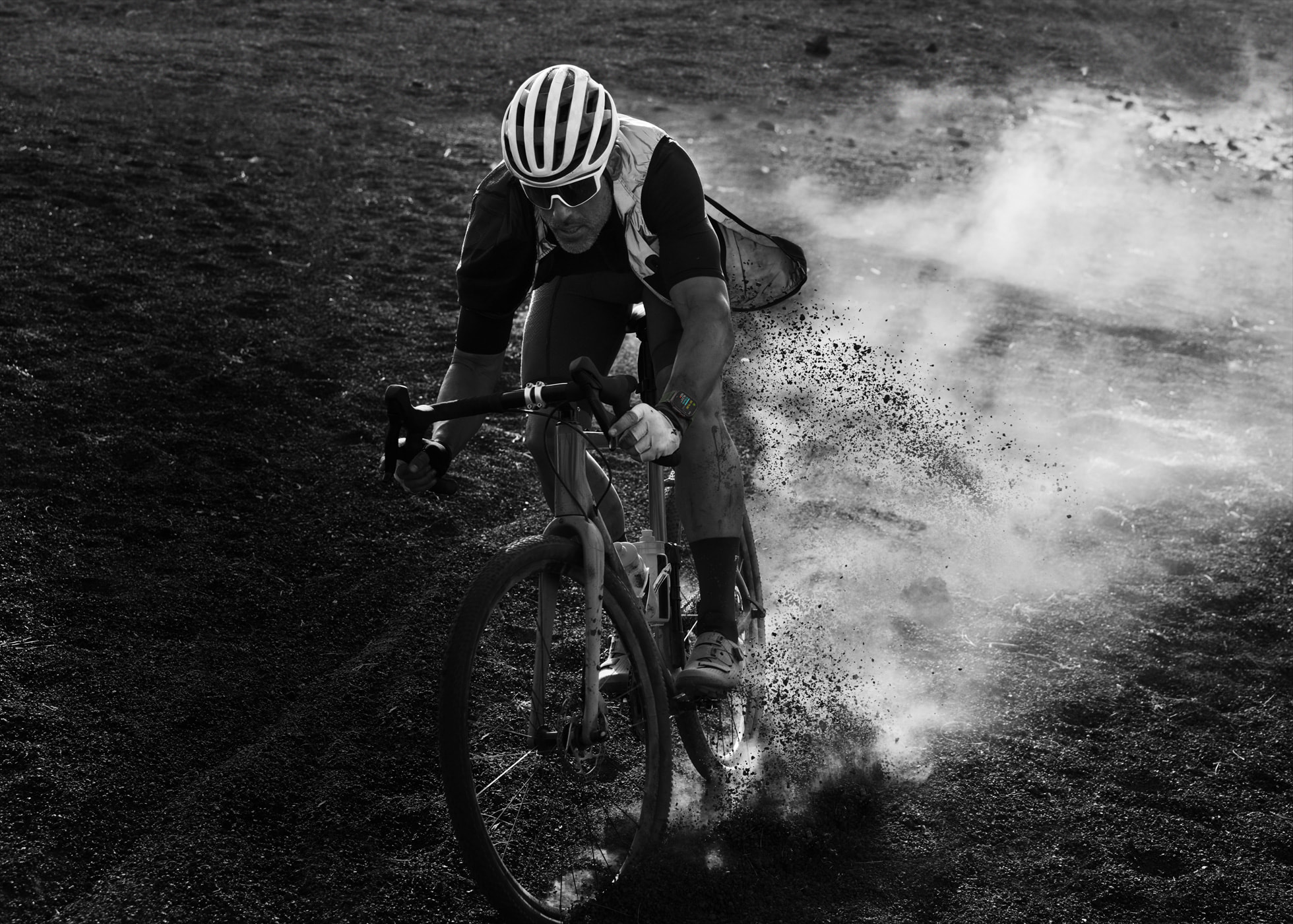 A cyclist wearing a helmet, sunglasses, and an Apple Watch Ultra 2 Black Titanium rides on a dirt path, kicking up a cloud of dust. The image is black and white, capturing the intense focus and effort of the rider as they maneuver through rough terrain.