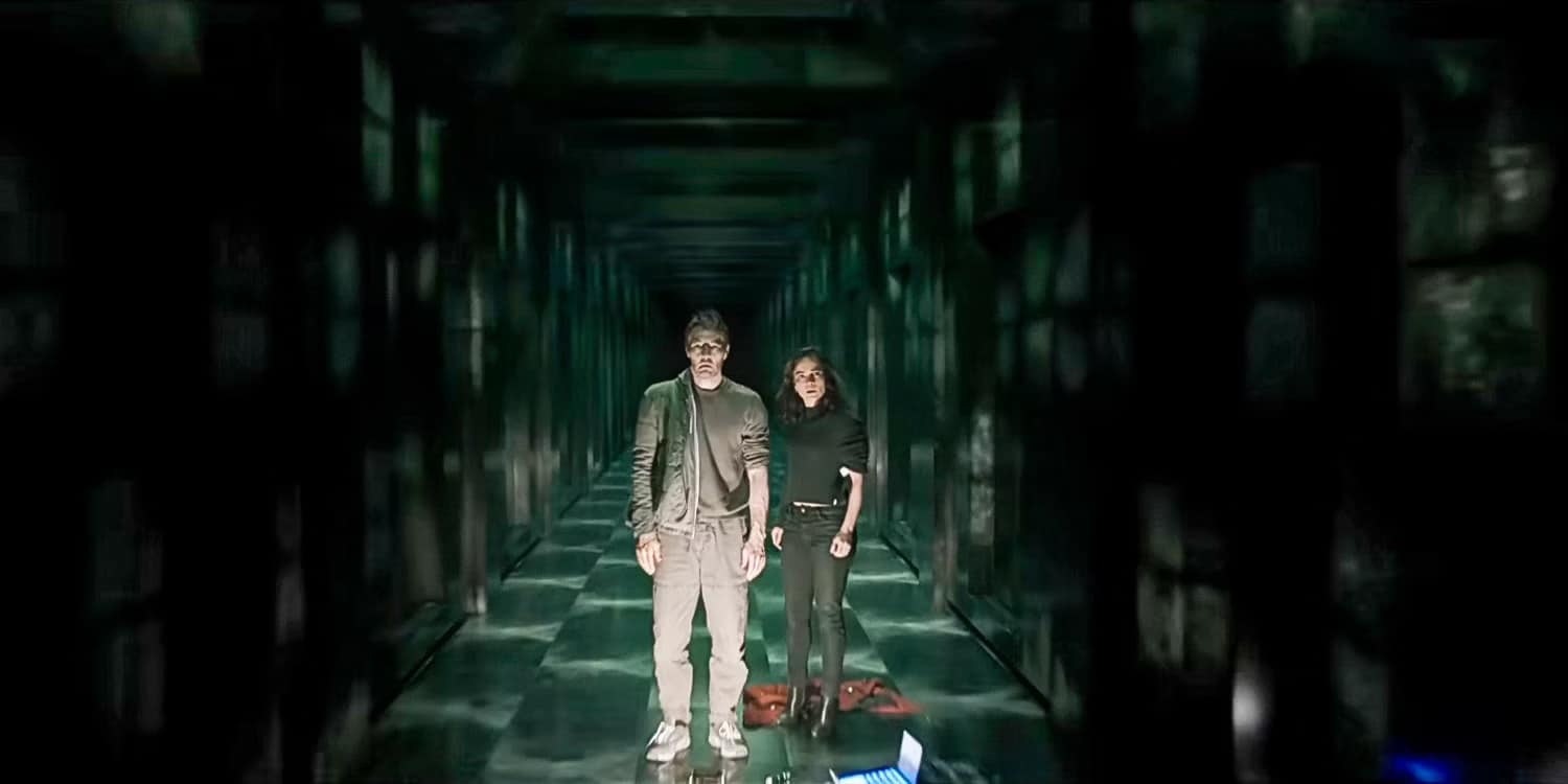 Dark Matter Series Season 1 | Apple TV+ | Two people stand in a dark, narrow hallway with a greenish, futuristic aura reminiscent of Dark Matter Season 2. One person wears a jacket and glasses, the other a black outfit. Both look forward with serious expressions. The setting has a sci-fi feel, illuminated by soft, grid-like lights.