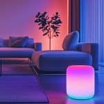 A modern living room with a minimalist design. A glowing smart speaker illuminates the foreground in gradient hues of pink and blue. The background features a stylish sofa, a coffee table, and a potted plant, all bathed in ambient lighting. AppleMagazine logo in the corner.