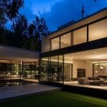 Modern two-story house with large glass windows and outdoor lighting, surrounded by trees and greenery. The interiors are well-lit, showing a spacious living area with contemporary furniture. The house features a sleek, minimalist design. A logo that reads AppleMagazine is visible in the bottom right corner.