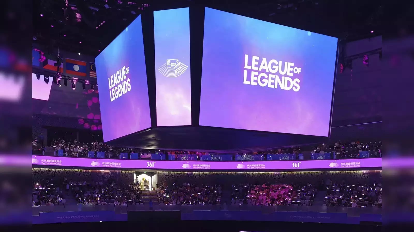 A large screen in an arena displays the "League of Legends" logo. The venue is filled with spectators, and purple-lit banners in various languages wrap around the upper level. The atmosphere suggests excitement and anticipation for a gaming event, promising a complete guide to the thrilling world of LoL Boosting Services.