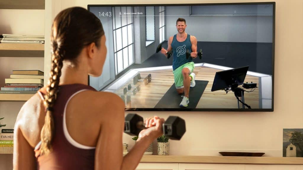 Apple Fitness vs Peloton: A person exercises at home, watching a fitness instructor on a TV screen. The instructor performs a lunge with one knee down, smiling and wearing athletic gear. The exercising person holds a dumbbell in one hand and is facing the TV. Fitness equipment is visible in the room as they explore Apple Fitness vs Peloton options.