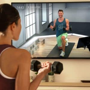 Apple Fitness vs Peloton: A person exercises at home, watching a fitness instructor on a TV screen. The instructor performs a lunge with one knee down, smiling and wearing athletic gear. The exercising person holds a dumbbell in one hand and is facing the TV. Fitness equipment is visible in the room as they explore Apple Fitness vs Peloton options.