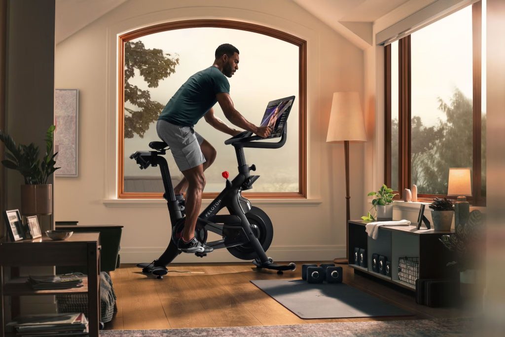 Apple Fitness vs Peloton: A person rides a stationary bike in a sunlit room with large windows, engaging with a screen showcasing an Apple Fitness vs Peloton comparison. The room is equipped with exercise gear, a yoga mat, dumbbells, and shelves. Natural light filters in, highlighting the cozy and organized workout space.