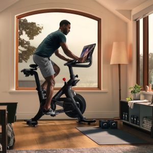 Apple Fitness vs Peloton: A person rides a stationary bike in a sunlit room with large windows, engaging with a screen showcasing an Apple Fitness vs Peloton comparison. The room is equipped with exercise gear, a yoga mat, dumbbells, and shelves. Natural light filters in, highlighting the cozy and organized workout space.