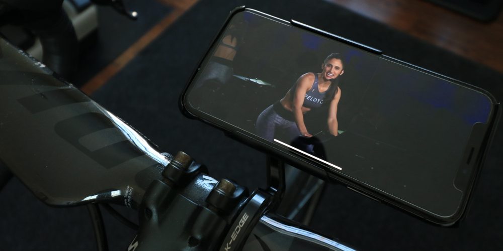 A smartphone mounted on an exercise bike's handlebars displays a fitness instructor leading a workout session. The instructor is smiling and engaging, wearing fitness attire. With discussions about Apple Fitness vs Peloton heating up, the bike's black handlebars and part of its frame are visible.